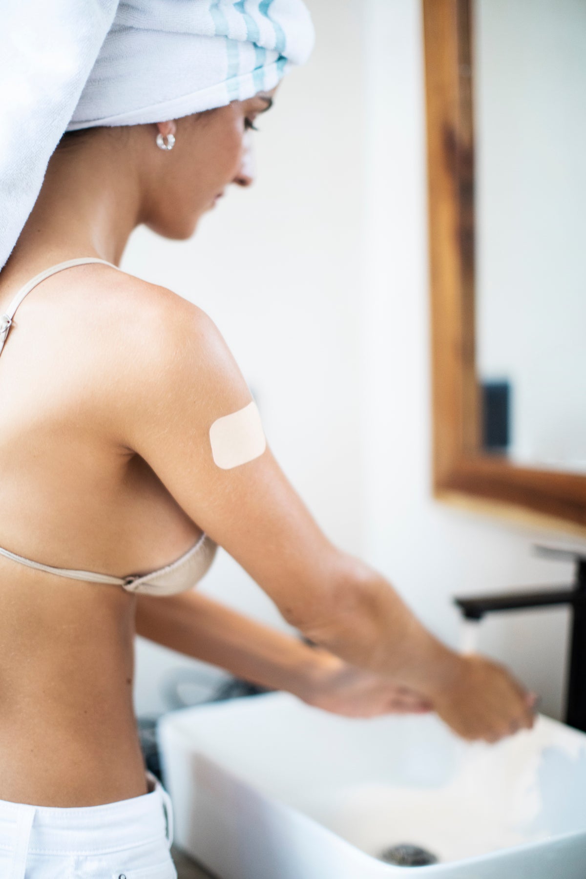 Woman with white bath towel on her head washing her hands in the bathroom with transdermal patch on her shoulder