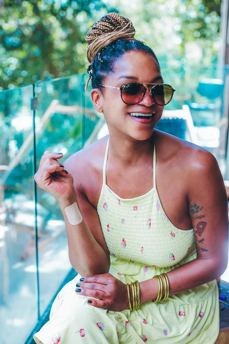 Smiling black woman in a yellow dress and sunglasses, wearing an Affix Patch on her wrist in a sunny, bright room