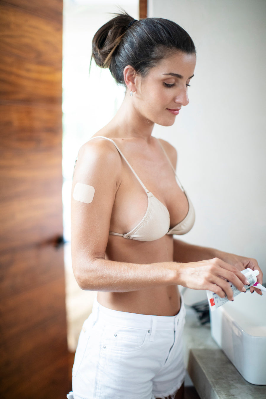 Attractive woman wearing a bra and white shorts brushing her teeth, wearing a transdermal patch on her shoulder