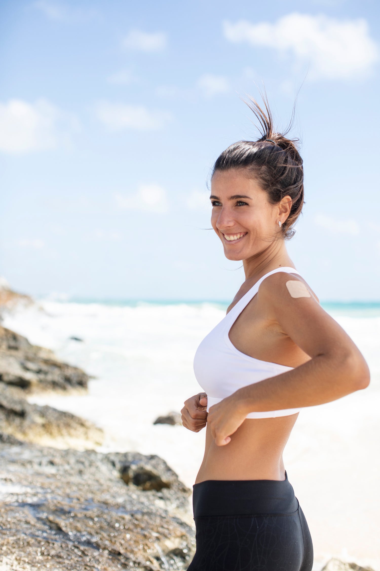 Fit, athletic young woman with brown hair, exercising on the beach, wearing an Affix Patch on her shoulder, looking joyful